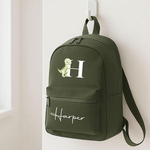 May include: Olive green backpack with a front pocket and a top handle. The backpack features a white letter "H" with a watercolor dinosaur illustration. The name "Harper" is written in white script on the front pocket.
