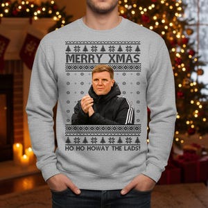 May include: Gray sweatshirt with a festive design. The text "MERRY XMAS" is displayed above a portrait. Below the portrait, the text "HO HO HOWAY THE LADS!" is printed. The design includes snowflakes and Christmas trees.