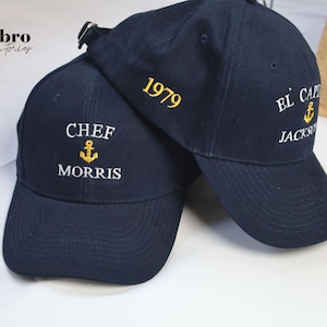 May include: Two navy blue baseball caps with embroidered white text and gold anchors. The cap on the left says "Chef Morris" and the cap on the right says "El Capitan Jackson 1979".