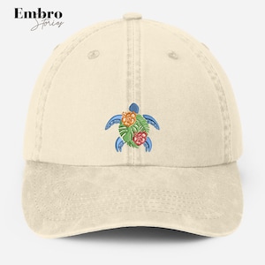 Pode incluir: Um boné de beisebol bege com uma tartaruga marinha bordada em azul, verde, laranja e vermelho com flores. Embro Stories está impresso na parte frontal do boné.
