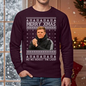 May include: A burgundy Christmas sweater with a festive design. The sweater features the text "MERRY XMAS" above an image of a man in a black jacket. Below the image is the text "HO HO HOWAY THE LADS!" with snowflake and Christmas tree patterns.