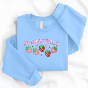 May include: A pale blue sweatshirt featuring the word "PEDIATRICS" in pink. The design includes embroidered strawberries, hearts, and flowers. The sweatshirt is made of a soft material.