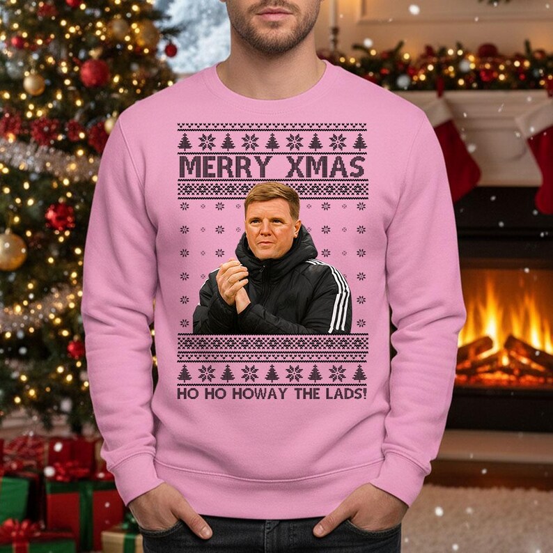 May include: Pink Christmas sweater with a festive design. The sweater features the text "MERRY XMAS" and "HO HO HOWAY THE LADS!". It also includes a portrait of a man. The design incorporates snowflakes and Christmas trees.