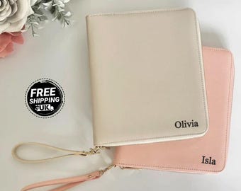 May include: Two personalized zip-around wristlet wallets. One is a light beige color with the name "Olivia" printed on it, and the other is a soft pink with the name "Isla". Both have gold-colored zippers and matching wrist straps. The background includes white flowers.