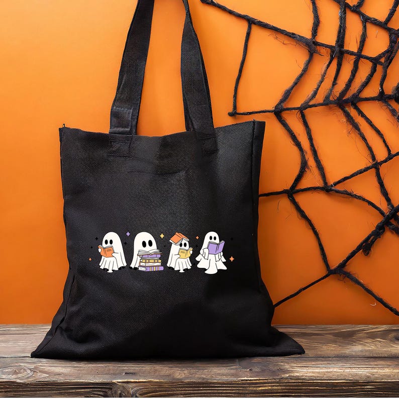 May include: Black tote bag featuring a Halloween-themed design. The graphic shows four cartoon ghosts, each holding or near books. The ghosts are white with black eyes and are set against a black background. The bag has black handles.