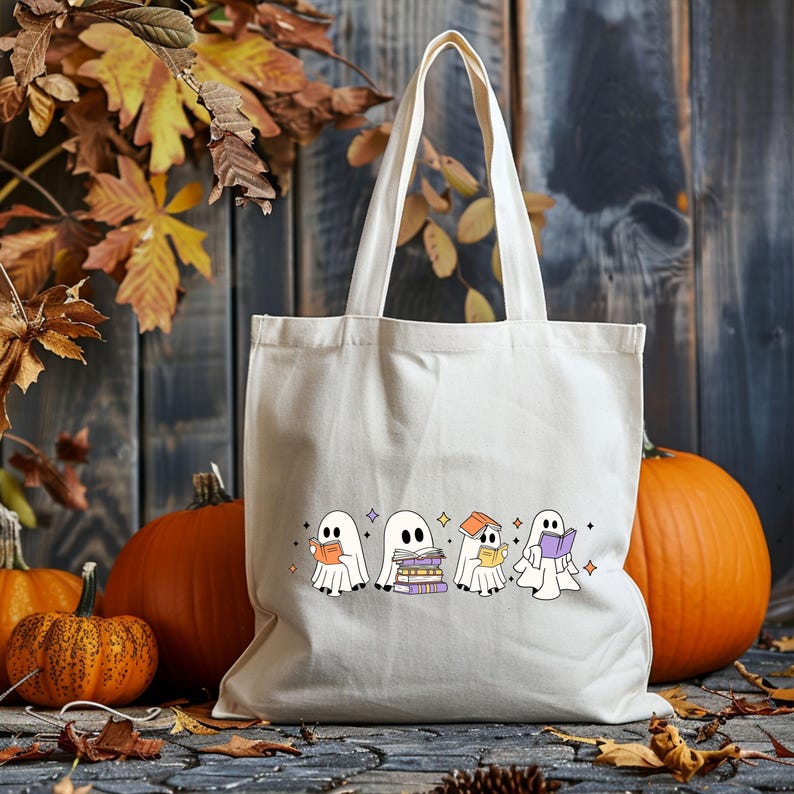 May include: Cream-colored tote bag with a Halloween-themed design. The graphic features four cartoon ghosts reading books, with pumpkins and fall foliage in the background. The bag has a rectangular shape and two handles.