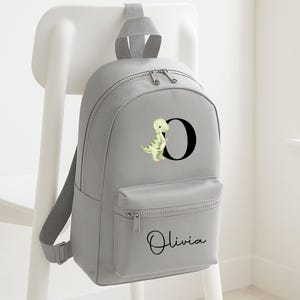 May include: A gray backpack with a front pocket and adjustable straps. The backpack features a black letter "O" with a green dinosaur illustration. The name "Olivia" is written in cursive below the pocket. The backpack is hanging on a white chair.