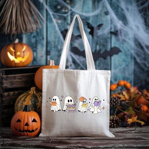 May include: A white canvas tote bag featuring a Halloween-themed design of cartoon ghosts reading books. The bag has long handles and is surrounded by carved pumpkins, gourds, and fall foliage, creating a festive autumn scene.