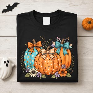 May include: A black t-shirt featuring a graphic of three pumpkins decorated with polka dots and bows in orange, blue, and teal. The pumpkins are surrounded by small flowers and leaves, creating a festive autumn-themed design on the front of the shirt.