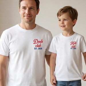 Pode incluir: Duas camisetas brancas, uma para um adulto e outra para uma criança. A camisa do adulto diz "Dads club EST. 2026" em escrita vermelha. A camisa da criança diz "Kids club EST. 2026" em escrita vermelha. Ambas as camisas são de manga curta.
