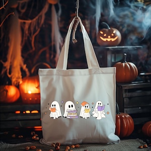 May include: Off-white tote bag featuring a row of cartoon ghosts reading books. The ghosts are white with black eyes and are holding books of various colors. The background includes pumpkins and Halloween decorations.