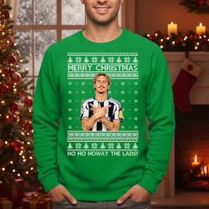 Puede incluir: Jersey navide&ntilde;o verde con el texto "MERRY CHRISTMAS" y "HO HO HOWAY THE LADS!". El jersey presenta un gr&aacute;fico de una persona con una camiseta a rayas blancas y negras. El jersey tiene un dise&ntilde;o festivo con copos de nieve y &aacute;rboles de Navidad.
