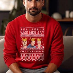 May include: Red crew neck sweatshirt with a festive design. The text "WISE MEN SAY" and "ONLY FOOLS RUSH IN" are displayed above and below a graphic of three men wearing crowns. The design includes snowflakes and Christmas trees.