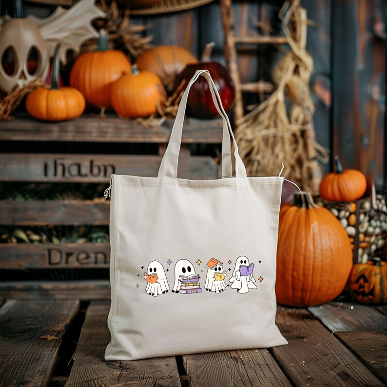 May include: A cream-colored tote bag featuring a line of cartoon ghosts reading books. The ghosts are wearing scarves and holding books. The background includes pumpkins, wooden crates, and fall foliage.