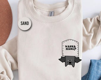 May include: Sand-colored sweatshirt with a bat graphic hanging from a pocket that says "Wanna Hang?". The sweatshirt is a neutral color and has a round neckline. The bat is black and gray.