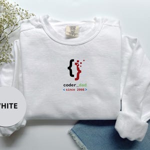May include: White crew neck sweatshirt with the text "coder_dad <since 2008>" in black, red, and green. The design features a stylized coding symbol. The sweatshirt is folded, with a tag visible on the collar.