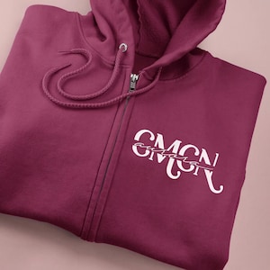 May include: A deep maroon zip-up hoodie featuring a hood with matching drawstrings. On the left chest, there is a white graphic design consisting of the large letters CMGN with the name aurora written in a cursive script across the center of the letters.