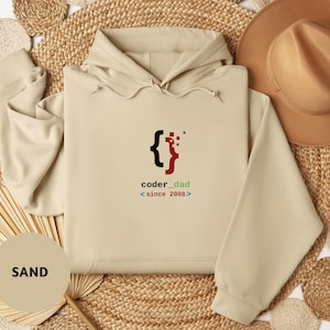May include: A sand-colored hoodie with a drawstring hood and long sleeves. The front features the text "coder_dad" with a graphic of curly braces and a stylized code symbol. The text "since 2008" is below the main text.