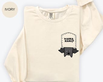 May include: Ivory-colored sweatshirt with a bat graphic. The bat is hanging from a pocket that says "WANNA HANG?" in black text. The sweatshirt has long sleeves and a crew neck.