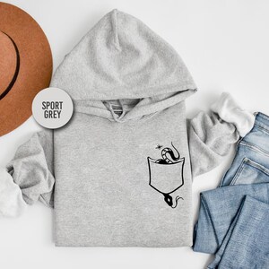 May include: A light gray hoodie with a hood and a kangaroo pocket. The pocket features a black graphic of a snake. The text "SPORT GREY" is printed on a circular label. The hoodie is paired with blue jeans and a brown hat.
