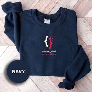 May include: Navy blue sweatshirt with the text "coder_dad" and the year "since 2008" in a coding-style font. The design includes a bracket symbol and a red and white graphic. A navy blue circle with the word "NAVY" is in the lower left corner.