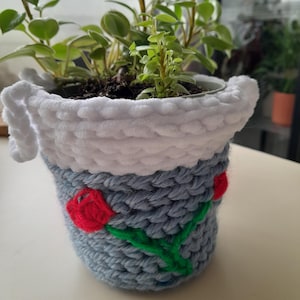 以下が含まれることがあります： 白の縁取りが付いた青い編み物の植木鉢。鉢には赤い刺繍のバラが2つと緑色の茎があります。