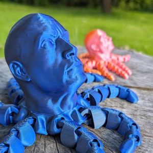 May include: A blue 3D printed octopus with a human face. The octopus has eight tentacles and is sitting on a wooden surface.