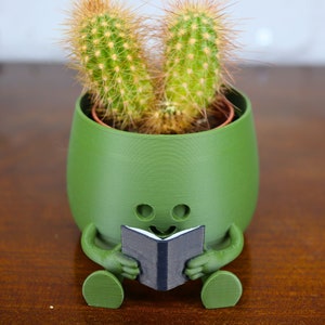 May include: A green ceramic planter shaped like a person sitting with legs crossed, holding a book. The planter has a smiling face and two eyes. A cactus plant is growing inside the planter.