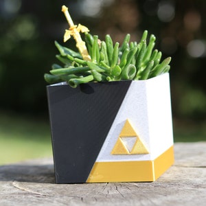 Puede incluir: Una maceta negra y blanca con un diseño triangular dorado y una espada dorada que sobresale de la parte superior. La maceta está llena de suculentas verdes.