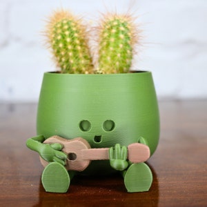 May include: A green ceramic planter shaped like a cartoon character playing a guitar. The planter has two cactus plants growing out of the top, resembling bunny ears.