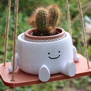 Może przedstawiać: Biała ceramiczna doniczka w kształcie uśmiechniętej postaci z dwoma nogami i ramionami, trzymająca małą roślinę kaktusową w brązowej doniczce. Doniczka jest zawieszona na sznurkowej huśtawce.