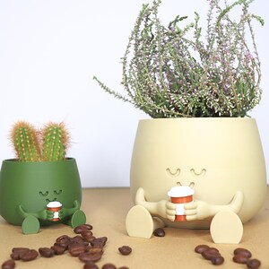 May include: Two green and beige ceramic planters shaped like cartoon characters with closed eyes and smiling mouths. The planters are sitting on a brown surface with coffee beans scattered around them. The planters are holding small coffee cups.
