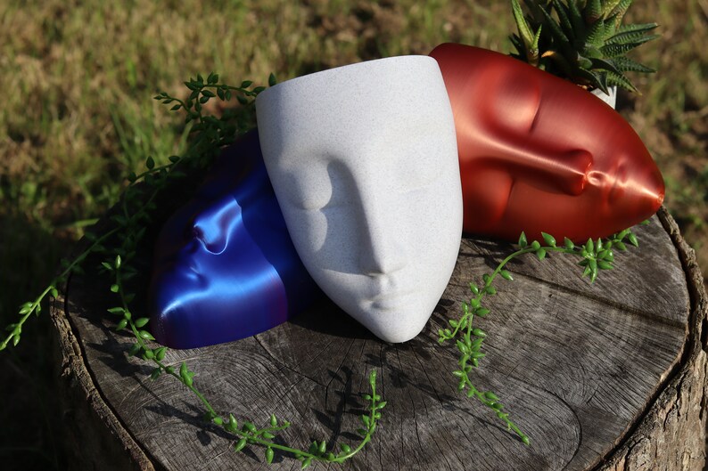 May include: Three ceramic face planters in blue, white and red. The planters are arranged on a tree stump with green foliage.
