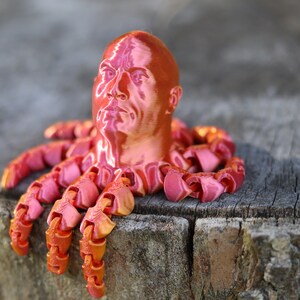 May include: A 3D printed sculpture of a human head with a red, metallic finish, attached to a set of octopus tentacles.