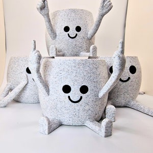 以下が含まれることがあります： 斑点模様の白いセラミック製のプランターが4つ。それぞれに黒く塗られた顔があり、2本の腕と2本の脚で人間のような形をしています。プランターは積み重ねられており、一番上のプランターの腕は空中に突き上げられています。