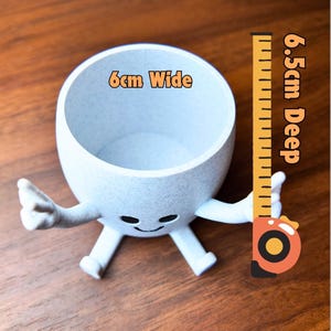 Puede incluir: Una maceta gris claro, caprichosa, con brazos, piernas y una cara sonriente. La maceta mide 6 cm de ancho y 6,5 cm de profundidad. La maceta est&aacute; sobre una superficie de madera.