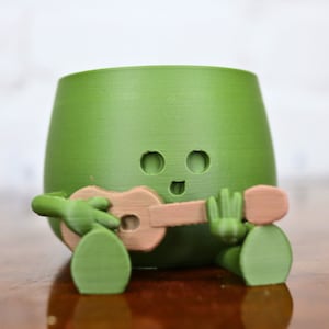 May include: A green ceramic planter shaped like a cartoon character playing a ukulele. The planter has a smiling face with two eyes and a mouth.