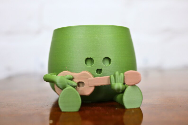 May include: A green ceramic planter shaped like a cartoon character playing a ukulele. The planter has a smiling face with two eyes and a mouth.