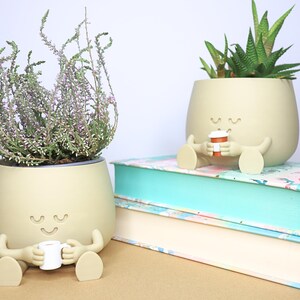 May include: Two beige ceramic planters shaped like a person with arms and legs, holding a small cup. The planter on the left has a plant with green leaves and pink flowers. The planter on the right has a succulent plant with green leaves. Both planters have a smiling face with closed eyes.