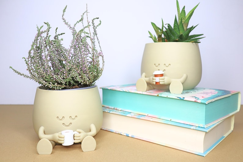 May include: Two beige ceramic planters shaped like a person with arms and legs, holding a small cup. The planter on the left has a plant with green leaves and pink flowers. The planter on the right has a succulent plant with green leaves. Both planters have a smiling face with closed eyes.