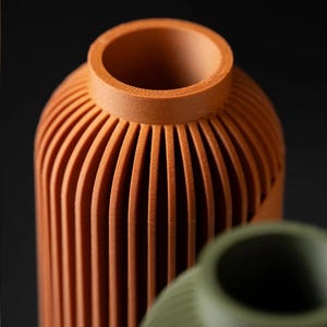 Peut inclure: Gros plan sur deux vases d&eacute;coratifs. Le vase avant est de couleur orange chaud avec des d&eacute;tails c&ocirc;tel&eacute;s verticaux. Le deuxi&egrave;me vase est de couleur verte sourde, &eacute;galement avec des d&eacute;tails c&ocirc;tel&eacute;s. Les deux vases ont une forme cylindrique et un dessus lisse et arrondi.