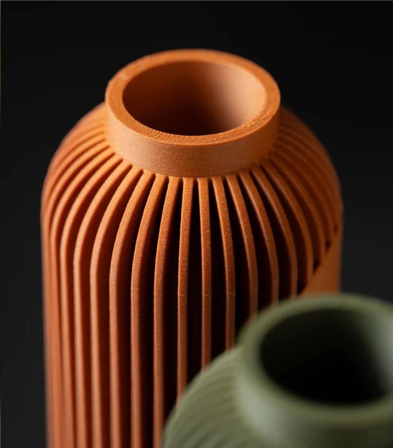 Peut inclure: Gros plan sur deux vases d&eacute;coratifs. Le vase avant est de couleur orange chaud avec des d&eacute;tails c&ocirc;tel&eacute;s verticaux. Le deuxi&egrave;me vase est de couleur verte sourde, &eacute;galement avec des d&eacute;tails c&ocirc;tel&eacute;s. Les deux vases ont une forme cylindrique et un dessus lisse et arrondi.
