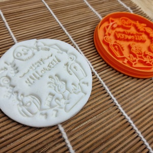May include: A white, round Halloween-themed cookie with embossed designs, including a skull, pumpkin, and the words "Happy Halloween." An orange cookie cutter with similar Halloween motifs sits nearby.