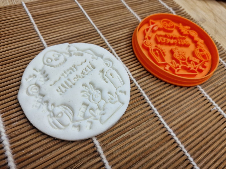 May include: A white, round Halloween-themed cookie with embossed designs, including a skull, pumpkin, and the words "Happy Halloween." An orange cookie cutter with similar Halloween motifs sits nearby.