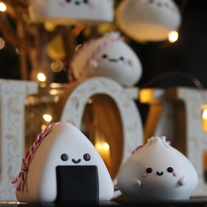 May include: Two cute, white, hand-crafted ornaments shaped like a rice ball and a dumpling. Both have black eyes and a smiling mouth. The rice ball has a black seaweed filling and a pink and white string tied around it. The ornaments are in front of a wooden "LOVE" sign with lights.