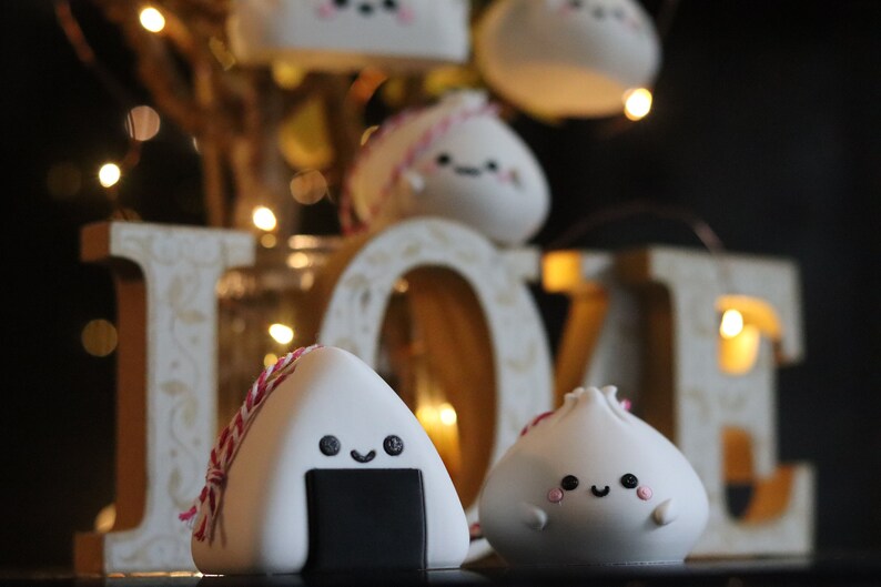 May include: Two cute, white, hand-crafted ornaments shaped like a rice ball and a dumpling. Both have black eyes and a smiling mouth. The rice ball has a black seaweed filling and a pink and white string tied around it. The ornaments are in front of a wooden "LOVE" sign with lights.