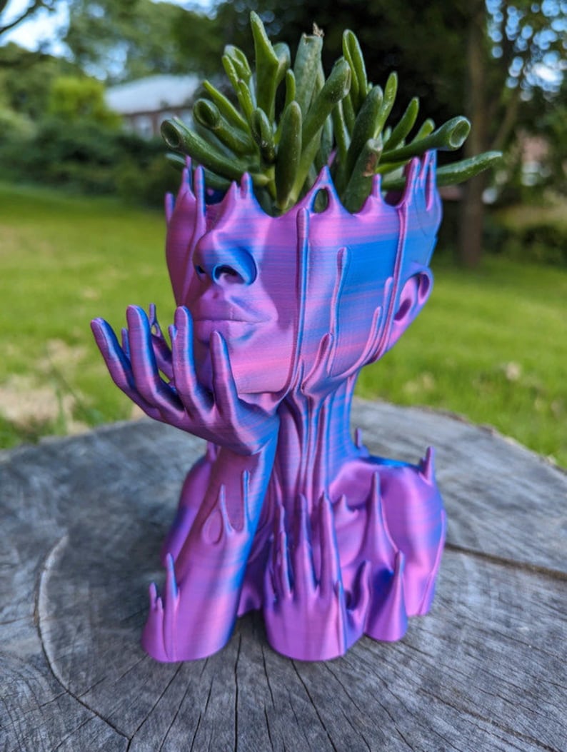 May include: A 3D printed planter shaped like a human head with dripping blue and purple paint. The planter has a small succulent plant growing out of the top.