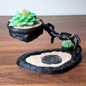 Op de afbeelding: Een zwarte, 3D-geprinte vetplantenpot met een kettingontwerp. De pot bevat een groene vetplant en staat boven een basis met zand en een kleine vetplant. Het ontwerp staat op een houten oppervlak.