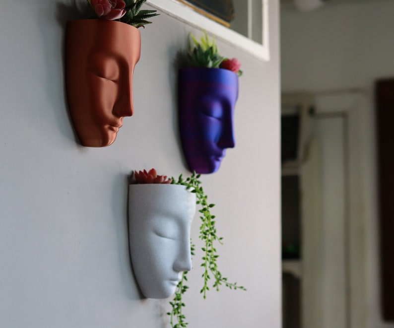 May include: Three wall-mounted planters shaped like faces, each with a different colour and a small succulent plant inside. The planters are made of ceramic and have a smooth, matte finish. The colours are a deep red, a vibrant purple, and a white.