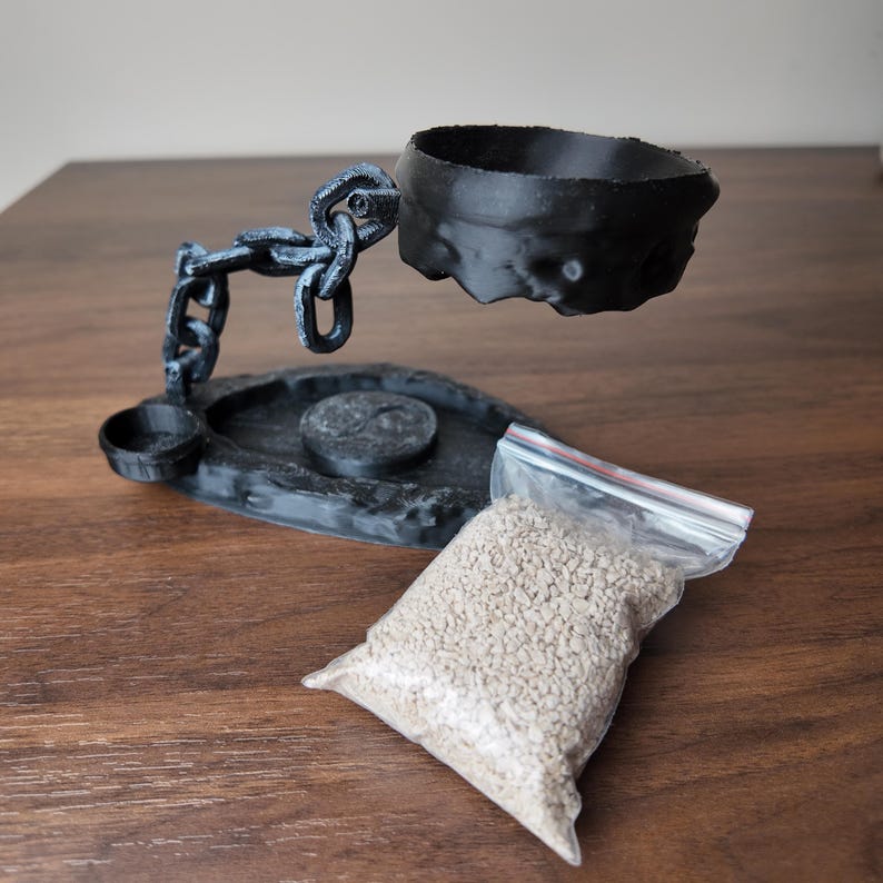 May include: A black, 3D-printed incense holder with a chain design supporting a bowl. A small dish and a bag of incense granules are also present. The holder is on a wooden surface.